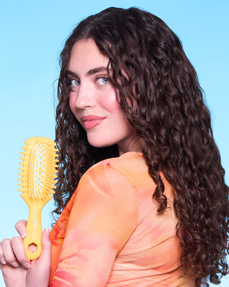 Woman with curly hair holding a yellow hairbrush against a light blue background