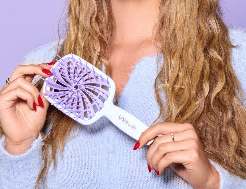 Woman wearing purple sunglasses and holding a white hair dryer with a purple attachment against a purple background