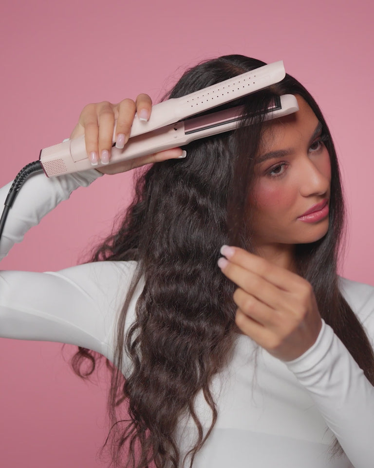 Woman with long, wavy hair holding a hair straightener against a pink background